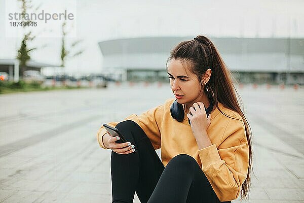 Außenaufnahme einer glücklichen jungen Frau mit Kopfhörern  die Musik auf ihrem Smartphone hört und lächelt  während sie auf der Straße in einem leeren Stadion sitzt  gekleidet in Freizeitkleidung