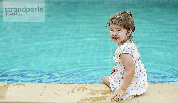 Rückenansicht eines entzückten Kindes in einem Kleid  das am Swimmingpool sitzt und lächelt  während es über die Schulter schaut