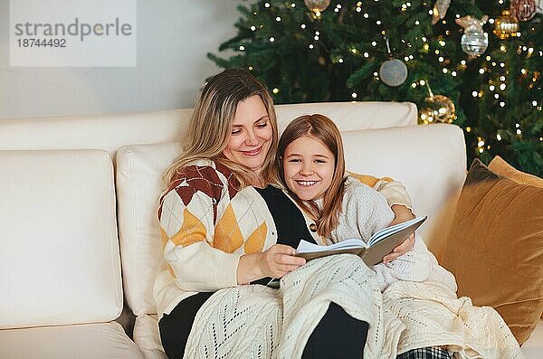 Glückliche Familie Großmutter und kleines Mädchen Tochter sitzen zusammen auf dem Sofa und Buch lesen  während Zeit zusammen zu Hause während der Weihnachtsferien im Winter  Umarmung und Abdeckung mit Wolle Plaid