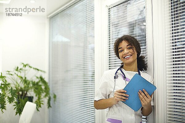 Glückliche afroamerikanische Ärztin in medizinischer Uniform mit Stethoskop um den Hals  die ein digitales Tablet hält und in die Kamera lächelt  während einer Arbeitsschicht in einer modernen Klinik
