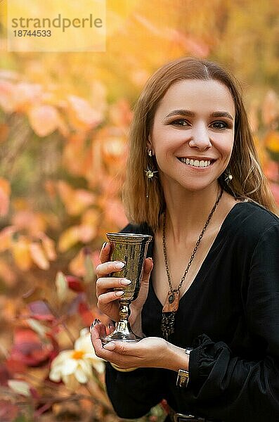 Junge fröhliche Frau in schwarzem Kleid hält Vintage Metall Glas in den Händen  während im Herbst Park mit bunten Blättern auf dem Hintergrund stehen  trinken Wein auf Picknick im Wald