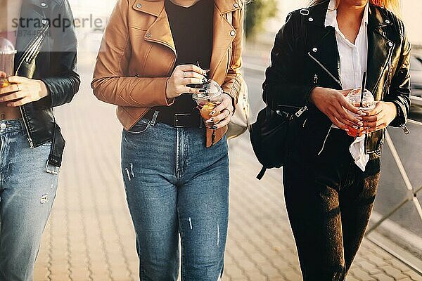 Glückliche junge Frauen mit Getränken zum Mitnehmen  die auf dem Bürgersteig spazieren gehen und an einem sonnigen Wochenendtag in der Stadt lächeln