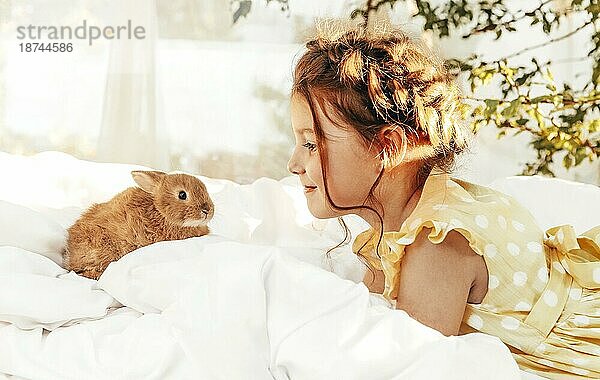 Kleines hübsches Mädchen im Sommerkleid sitzt draußen auf dem Bett mit Kaninchen  niedliche Kind Blick auf Hasen mit leichten Lächeln genießen Zeit mit ihren Lieblings Tiere