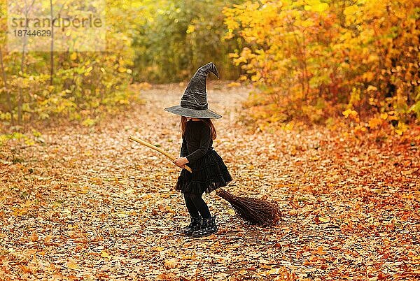 Nette glückliche kleine rothaarige Mädchen in Hexenkostüm sitzt auf Besen über Herbst Wald Hintergrund mit Kopie Raum  hält ihren Hut mit den Händen. Urlaub von Halloween Feier Konzept