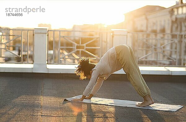Junge  sportliche  fröhliche  gemischtrassige Frau in Sportkleidung macht Dehnungsübungen  während sie auf einer Yogamatte auf dem Hausdach am frühen Morgen sitzt  fitte Frau  die bei Sonnenaufgang trainiert