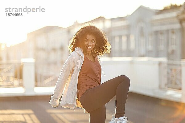 Junge glückliche sportliche afroamerikanische Frau  die auf dem Dach Sport treibt und in die Kamera lächelt  fitte gemischtrassige Frau  die während des morgendlichen Workouts auf einer Terrasse mit Stadtlandschaft im Hintergrund steht
