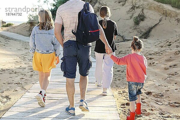 Foto von hinten von glücklichen Familie zu Fuß auf hölzernen Weg auf sonnigen Sandstrand  Vater und kleine Tochter Hand in Hand gehen mit anderen Schwestern in Richtung Meeresufer. Reisen und Sommerurlaub Konzept