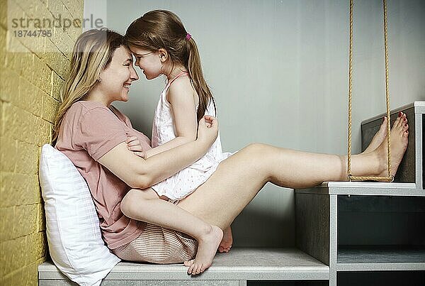 Junge liebevolle Mutter umarmt mit ihrer süßen kleinen Tochter  während sie zusammen auf der Treppe zu Hause sitzen  Mutter und kleines Kind verbringen Zeit zusammen am Wochenende  spielen drinnen. Familie und Elternschaft