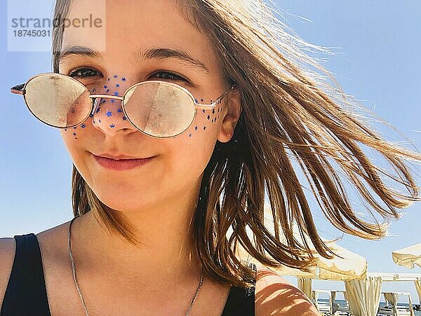 Teenagermädchen mit modischer Brille und bemaltem Gesicht entspannt sich am Strand und genießt den Sommerurlaub