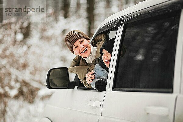 Fröhliche erwachsene Frau und kleiner Junge in Oberbekleidung  die in die Kamera schauen  während sie aus dem Fahrzeugfenster schauen  vor einem unscharfen Hintergrund eines Winterwaldes