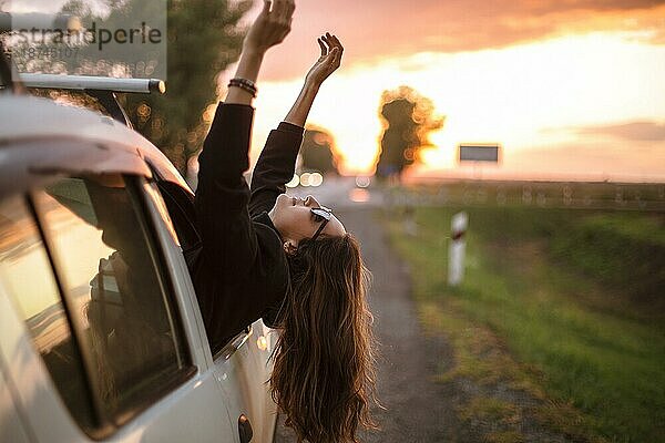 Junge sorglose Frau Reisende streckt sich aus dem Autofenster während der Fahrt durch die Landschaft auf schönen Sommerabend bei Sonnenuntergang  glücklich weiblichen Reisen mit dem Auto in der Natur. Reisen und Roadtrip