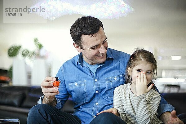 Fröhlich Vater spielt mit kleinen Tochter auf der Couch sitzen und helfen  ihre Nägel mit hellen Lack zu malen zusammen spielen. Junge Brünette Vater mit seiner süßen kleinen Tochter zu Hause