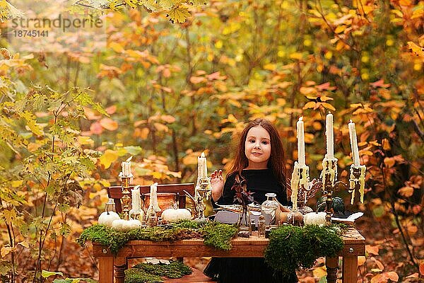 Kleine bezaubernde Mädchen in schwarzen Hexenkostüm spielen im Herbst Wald  Kind  das Spaß an Holztisch mit Moos  Kürbisse und verschiedene Dekorationen  viele brennende Kerzen. Halloween und Erntedank