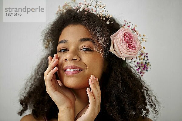 Close up Frühling Porträt der wunderschönen lächelnden afrikanisch amerikanischen jungen Frau mit frischen rosa Rose Blume in lockiges Haar Blick in die Kamera  vorgrauem Hintergrund. Natürliche Schönheit der afrikanischen Frauen
