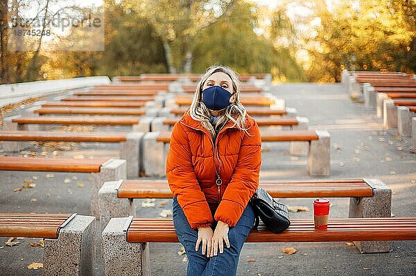 Blonde Frau in modischer Oberbekleidung und Stoffmaske schaut in die Kamera  während sie auf den Bänken im Park während der Pandemie sitzt