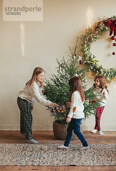 Glückliche vier kleine Geschwister  drei Schwestern in warmen Strickpullovern versammeln sich um einen Tannenbaum im Wohnzimmer und dekorieren ihn mit Spielzeug und glänzenden Kugeln. Familie und Weihnachten Konzept