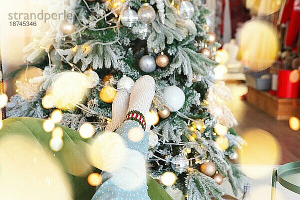 Weihnachten und Neujahr Hintergrund mit weiblichen Füssen in warmen gestrickten Socken gegen Weihnachtsbaum mit gelben Glühbirnen Girlande geschmückt  selektiven Fokus. Frau entspannt zu Hause in der Weihnachtszeit  beschnittenes Foto