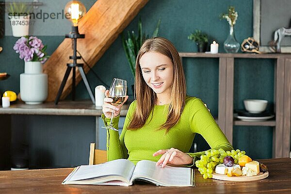 Junge Frau liest eine interessante Zeitschrift an einem Holztisch mit Obst und Wein an einem Wochenende zu Hause