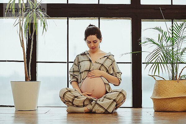 Junge ruhige schwangere Frau im Pyjama in voller Länge  die ihren Bauch hält  während sie im Lotussitz vor einem Panoramafenster sitzt. Die werdende Mutter berührt sanft ihren Bauch. Meditation während der Schwangerschaft