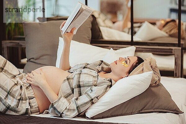 Junge  attraktive  glückliche schwangere Frau in Hauskleidung  die ein Buch liest und ihren Bauch berührt  während sie sich zu Hause auf der Couch ausruht. Die werdende Mutter trägt einen karierten Pyjama und genießt ihre Freizeit während der Schwangerschaft