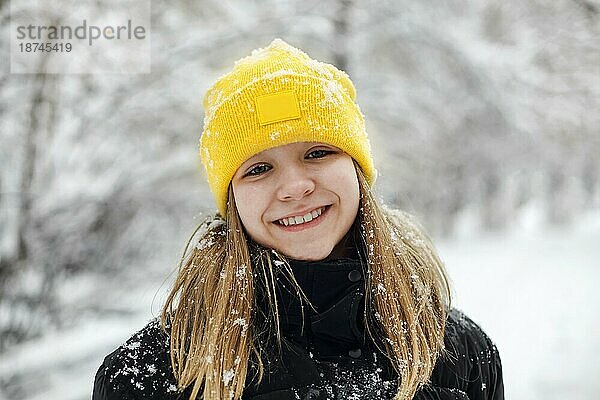Close up Porträt von lustigen glücklichen Teenagermädchen in gelben Strickmütze spielen mit Schnee im Freien  Kind hält großen Schneeball in der Hand und zeigt es der Kamera. Winter Schnee Spaß für Kinder in der Natur