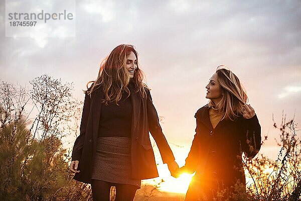 Zwei glückliche Frauen  die sich an den Händen halten  spazieren durch ein herbstliches Feld bei Sonnenuntergang und lächeln  sorglose beste Freundinnen  die einen Abendspaziergang auf einer Wiese auf dem Lande genießen