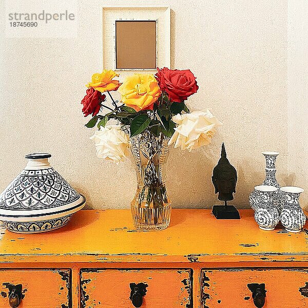 Schöner Strauß frischer mehrfarbiger Rosenblüten in Glasvase auf gelbem Vintage Tisch mit Buddhakopf und verschieden großen antiken Keramikvasen  ohne Menschen. Warm und gemütlich nach Hause Dekor