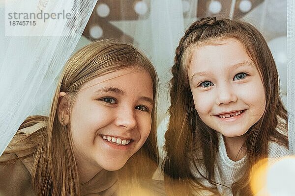 Gemütliche inländischen Porträt von zwei glücklichen lächelnden kleinen Mädchen Schwestern umarmt  während die Freizeit zusammen zu Hause zu verbringen  sitzen auf dem Bett in der Nähe geschmückten Weihnachtsbaum. Familie und Kinder Konzept