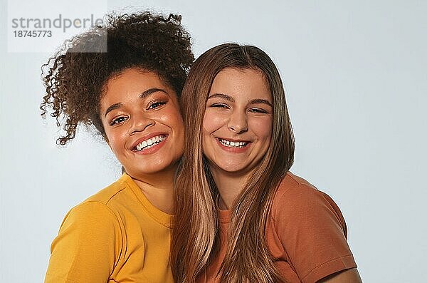 Positive multirassische Teenagermädchen interrassische beste Freundinnen  die dicht beieinander stehen  sich sanft Wange an Wange kuscheln und in die Kamera schauen und dabei ehrlich und strahlend vor dem Hintergrund einer weißen Wand lächeln