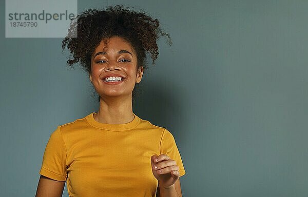 Schöne glückliche dunkelhäutige afrikanische amerikanische ethnische Frau im gelben TShirt die die Hand zur Begrüßung hebt  während sie mit einem angenehmen Lächeln in die Kamera blickt  posiert vor einem grauen  grünen Studiohintergrund