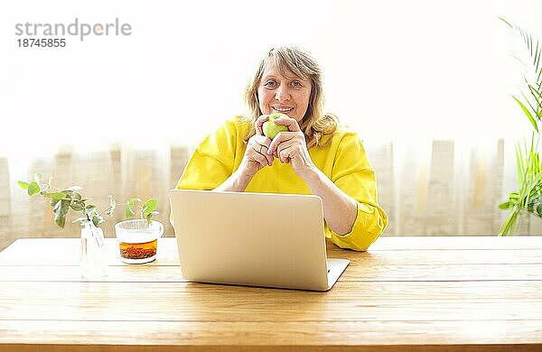 Positive Frau mittleren Alters in Freizeitkleidung sitzt am Holztisch mit einer Tasse Tee und einem Apfel und surft auf dem Laptop  während sie den Morgen zu Hause verbringt