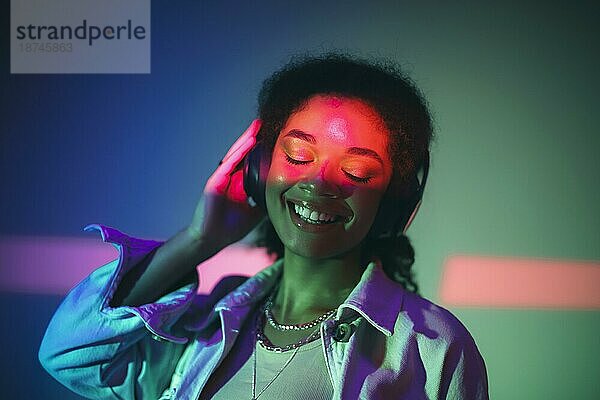 Junge glückliche afroamerikanische Frau mit schwarzen Kopfhörern  die Musik hört und hochwertigen Sound genießt  während sie unter lila Neonlicht im Studio steht und glücklich lächelt. Musikalische Ausrüstung