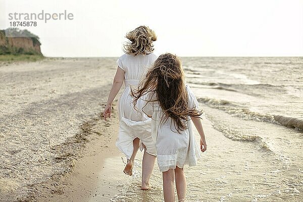 Ganzkörper fröhliche Frau in weißem Kleid mit kleinem Mädchen läuft am Strand in der Nähe winken Meer am Morgen