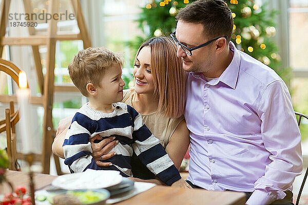 Erwachsener Mann und Frau umarmen glücklichen Sohn und Frau  während sie am Esstisch in einem gemütlichen Raum zu Hause während der Urlaubsfeier sitzen