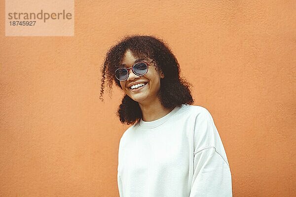 Junge Frau afrikanischer Abstammung mit stilvoller Sonnenbrille  mit lockigem Haar  das zu einem hohen Pferdeschwanz gebunden ist  schaut weg  während sie breit lächelt und gerade  perfekte Zähne zeigt  und vor einem orangefarbenen Wandhintergrund posiert