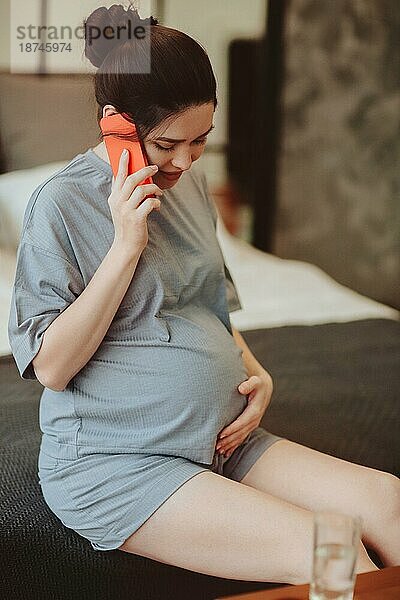 Junge lächelnde schwangere Frau  die ihren Bauch berührt und mit einem Freund am Handy spricht  während sie Zeit zu Hause verbringt  werdende Mutter in Hauskleidung  die auf dem Bett sitzt  ihren Bauch streichelt und über ihr Smartphone telefoniert