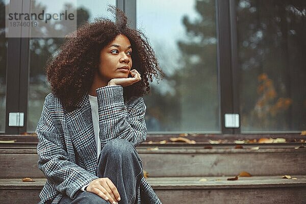 Outdoor Herbst Porträt der jungen nachdenklich afrikanischen amerikanischen Frau zur Seite schauen sitzen auf alten hölzernen Haus Treppe  nachdenklich lockiges Mädchen verbringen Zeit im Freien auf Herbst Tag  denken über das Leben