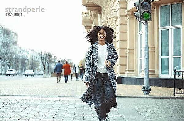 Junge stilvolle positive afroamerikanische Frau überqueren Zebrastreifen und lächelnd in die Kamera beim Gehen in der Stadt im Herbst Tag. Glückliches schwarzes Mädchen auf dem Zebrastreifen im Freien. Menschen auf den Straßen der Stadt