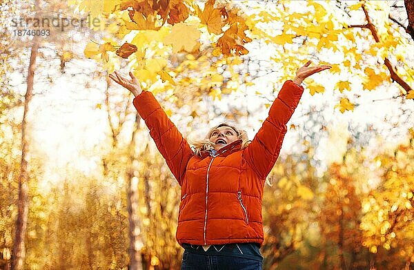 Zufriedene Frau in Oberbekleidung lächelnd mit trockenen Blättern an einem sonnigen Wochenendtag im Herbstpark