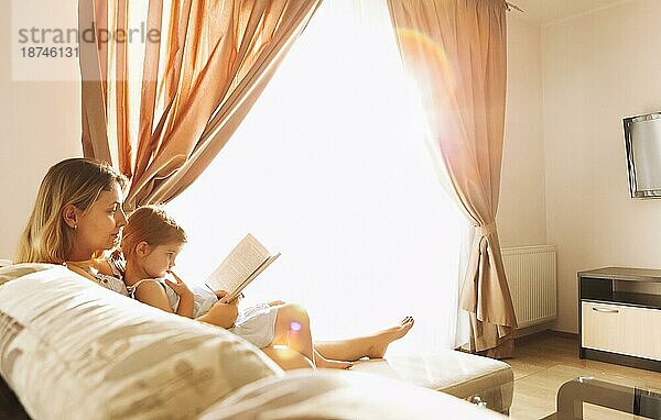 Entspannte Mutter und kleine Tochter sitzen zusammen auf einem gemütlichen Sofa im Wohnzimmer mit einem Buch in der Hand. Die Mutter und das süße Mädchen verbringen ihre Zeit sinnvoll mit Lesen und Lernen und genießen das Wochenende zu Hause