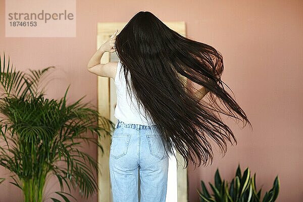 Rückenansicht einer schlanken jungen Brünetten mit langen Haaren in Freizeitkleidung  die neben einem Spiegel und Topfpflanzen an einer rosa Wand steht