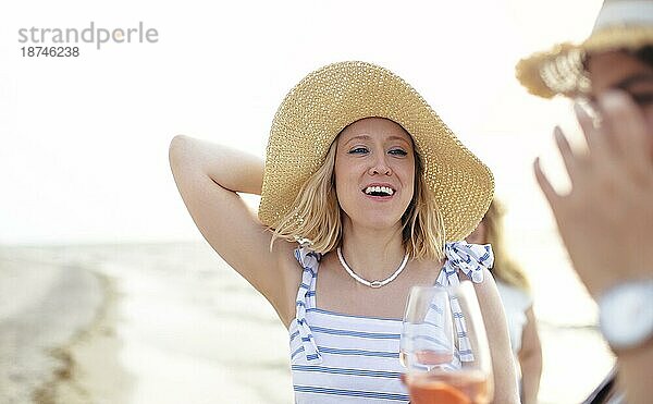 Fröhliche adultes Weibchen in Sommerkleidung lächelt und schaut weg  während sie am Sandstrand in der Nähe des Meeres Wein genießt