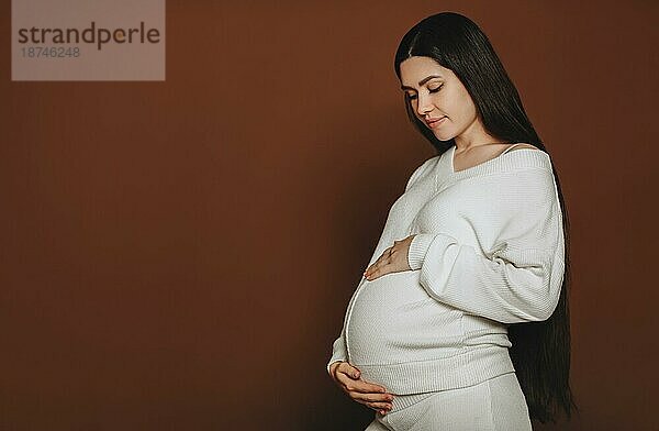 Junge glückliche schwangere Frau mit langen braunen Haaren  die den Bauch berührt und mit dem ungeborenen Baby spricht  während sie vor einem dunklen Studiohintergrund steht. Die werdende Mutter in weißer Kleidung streichelt den Bauch. Schwangerschaft