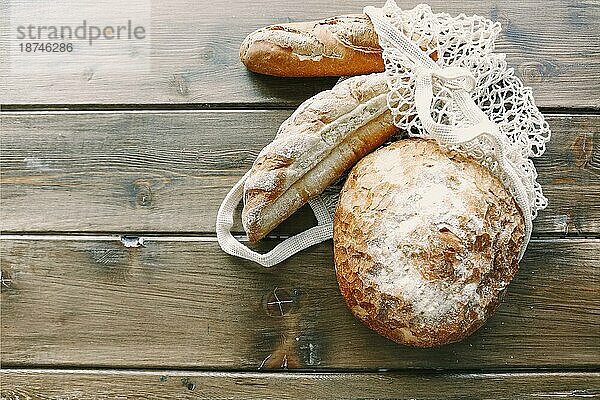 Draufsicht auf drei Laibe von verschiedenen Sorten von frisch gekauftem Brot liegt in weißen Lebensmittelgeschäft Netztasche auf Holz Vintage rustikalen Stil Tischoberfläche. Natürliche  organische und hausgemachte Lebensmittel Konzept