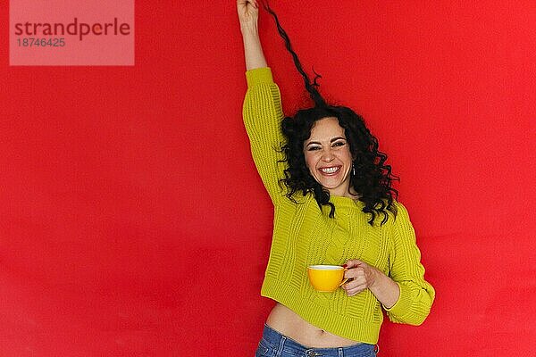 Verspielte Stimmung. Studioporträt von glücklichen überglücklich junge Frau mit Teetasse in den Händen spielen mit ihren lockigen Haaren und lächelnd fröhlich in die Kamera  vorrotem Hintergrund mit Kopie Raum für Text