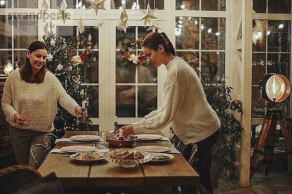Zwei junge Frauen decken festlichen Weihnachtstisch zusammen zu Hause  glückliche Mutter und Tochter Vorbereitung für Weihnachtsfeier in großen gemütlichen rustikalen Bauernhaus. Urlaub Traditionen Konzept