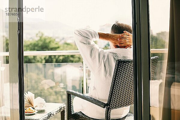 Rückenansicht eines anonymen Mannes  der die Hände hinter dem Kopf hält und auf einem Stuhl chillt  während er seine Zeit auf dem Balkon an einem sonnigen Sommertag verbringt