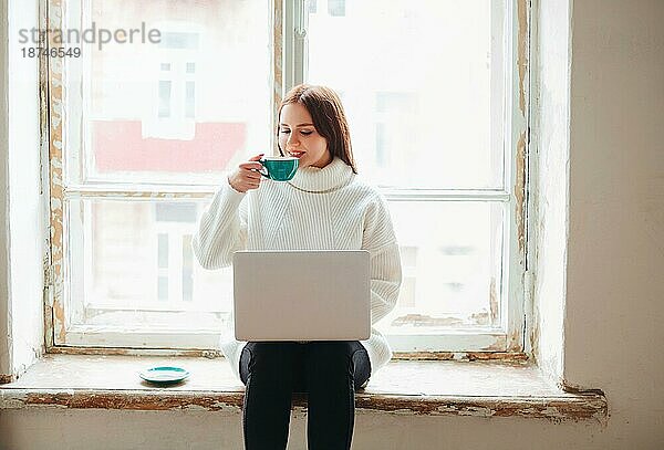 Fröhliche junge Frau genießt frischen Kaffee und surft auf ihrem Netbook  während sie auf der schäbigen Fensterbank vor dem Fenster sitzt