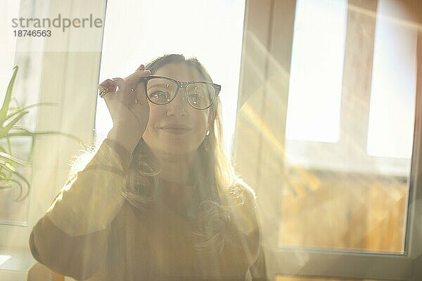 Fröhlich lächelnde Frau mittleren Alters  die eine Brille hält  durch das Glas schaut und lächelt  während sie in einem Raum sitzt  in dem die Sonne durch das Fenster ins Haus fällt  zufriedene blonde Frau  die gerne von zu Hause aus arbeitet