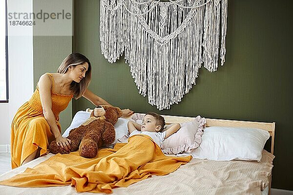 Gute Nacht Geschichte. Liebevolle Mutter setzen Sohn zu Bett für den Tag schlafen  Mutter sitzt neben ihrem Kind mit Teddybär Spielzeug im Bett zu Hause liegen. Lächelnde Frau liest Märchen für kleinen Jungen